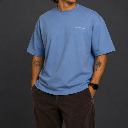 A man wearing a light blue crewneck t-shirt with a small white 'SPEED SHOP' logo on the left chest, set against a solid sage green background.