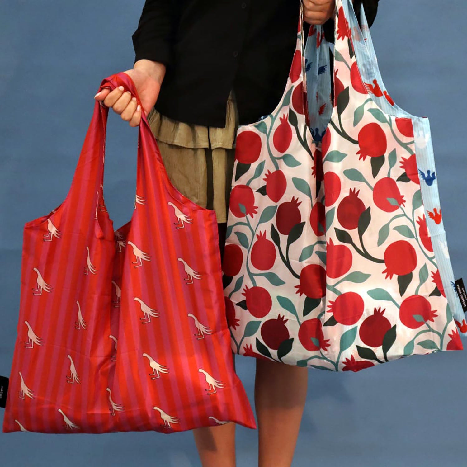 A person holds two reusable tote bags against a blue backdrop. One bag is bright red with a small white bird print, and the other is white with a bold red pomegranate and green leaf pattern, plus a light blue side panel.