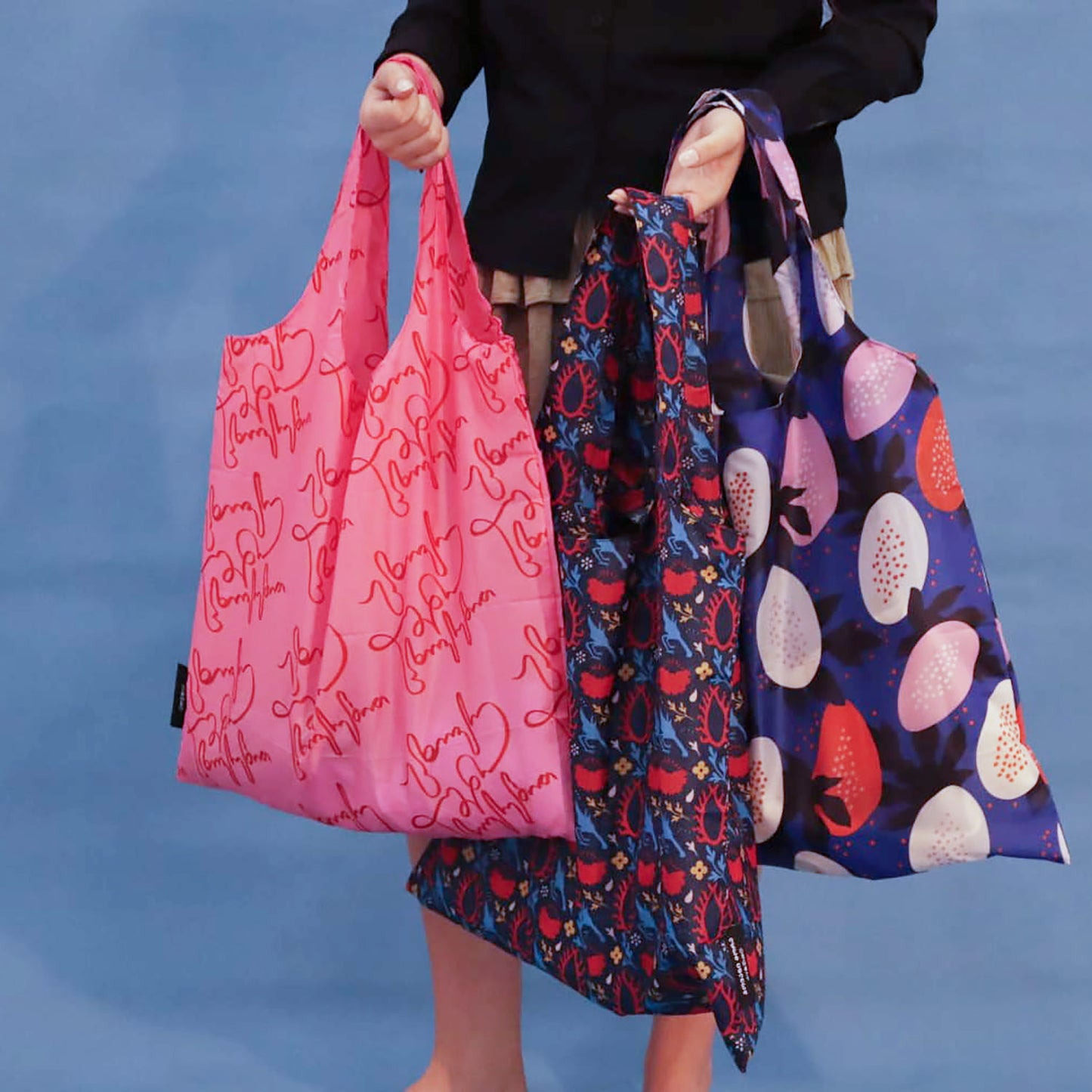 Three reusable tote bags held by a person against a blue backdrop: a bright pink bag with red script print, a dark blue patterned bag with red and blue motifs, and a dark blue bag with large pink, white, and red fruit-like prints.