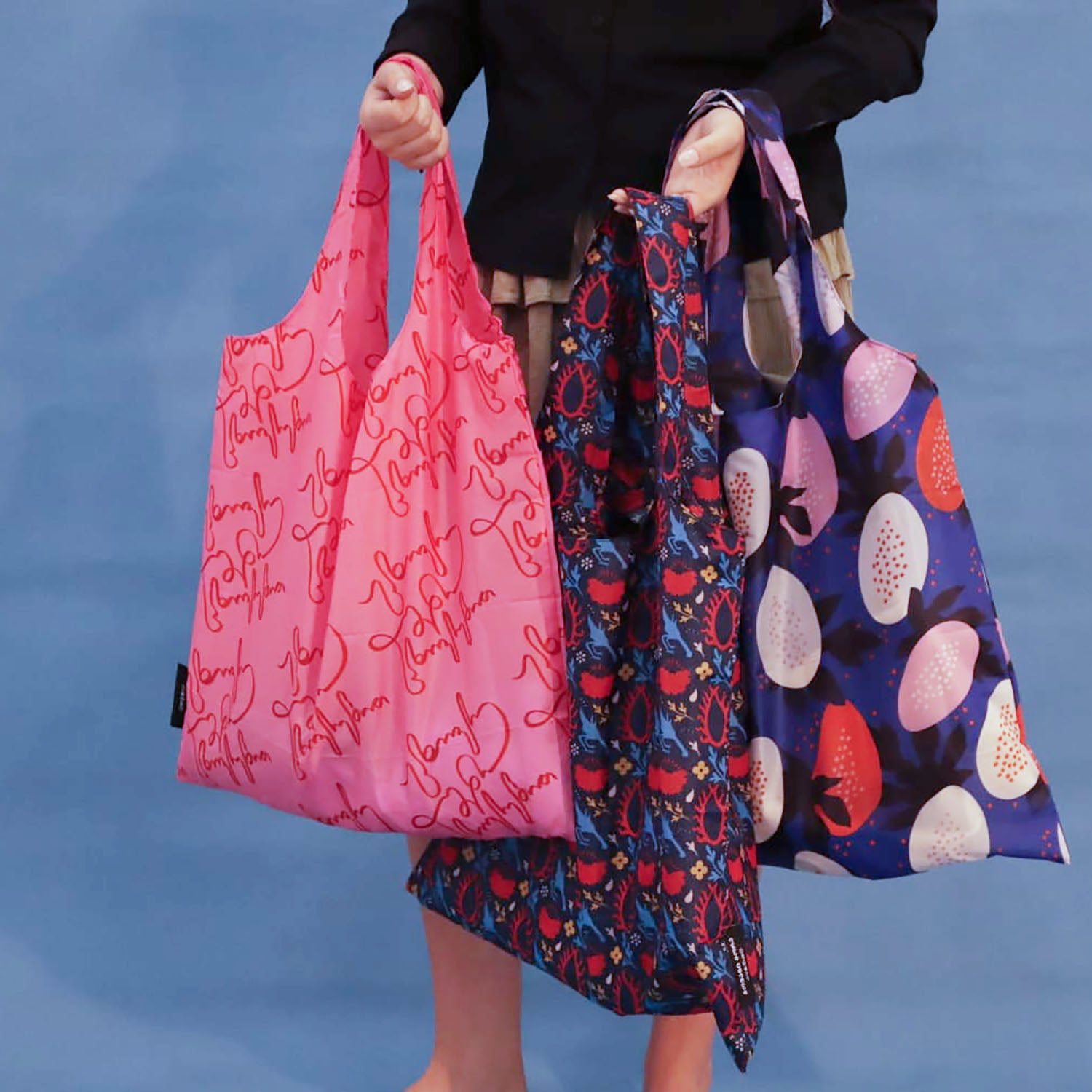 Three reusable tote bags held by a person against a blue backdrop: a bright pink bag with red script print, a dark blue patterned bag with red and blue motifs, and a dark blue bag with large pink, white, and red fruit-like prints.