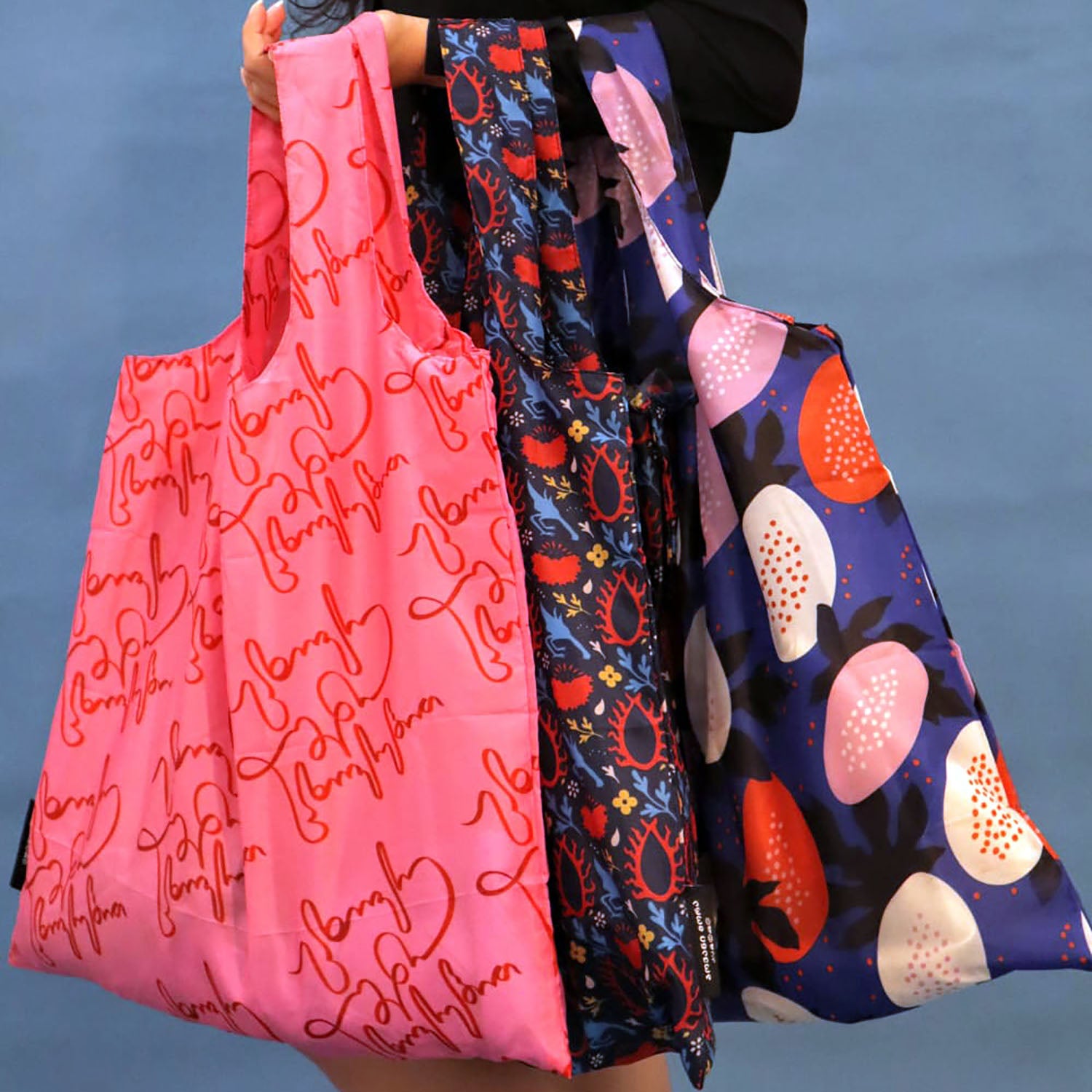 Three reusable tote bags held by a person against a blue wall. The left bag is bright pink with red script text, the middle bag has a dark blue and red floral pattern, and the right bag is dark blue with large pink, orange, and white fruit-like motifs.