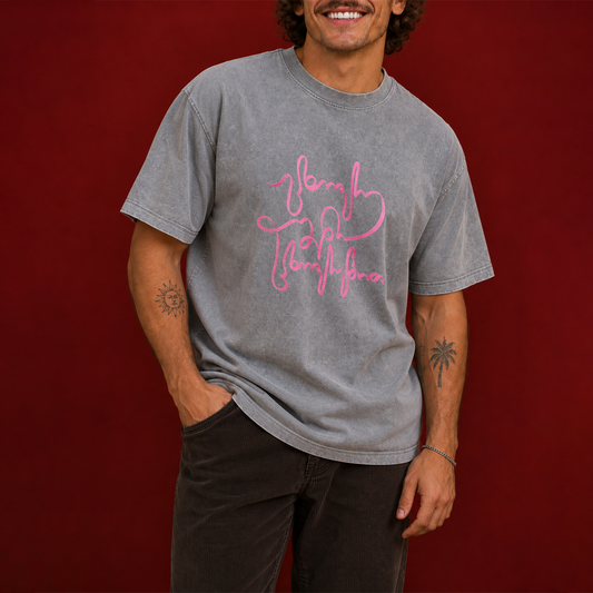 A man wearing a gray mineral-wash t-shirt with a pink script graphic on the front, paired with dark pants, standing against a solid dark red background.