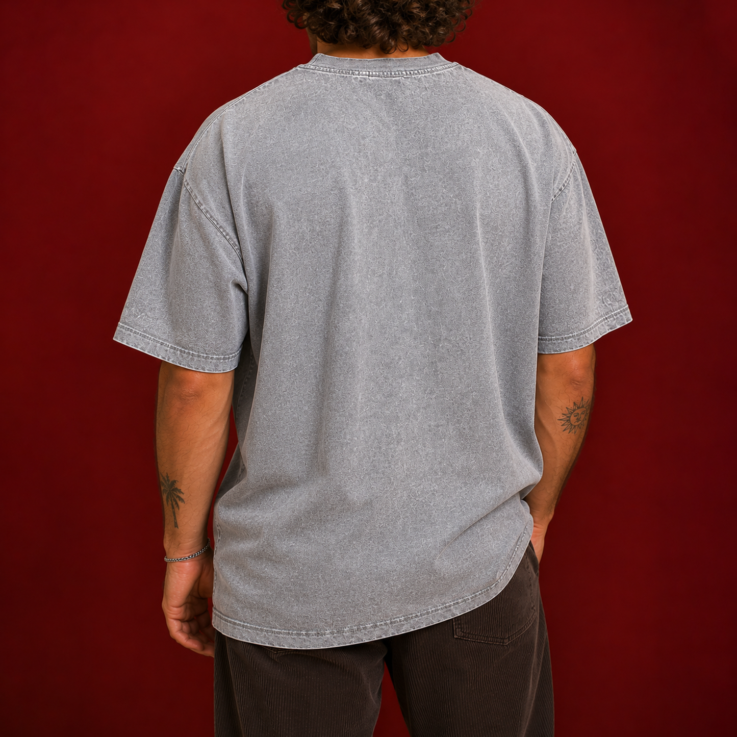 A person shown from the back wearing an oversized light gray washed t-shirt and dark brown pants, standing against a solid dark red background.