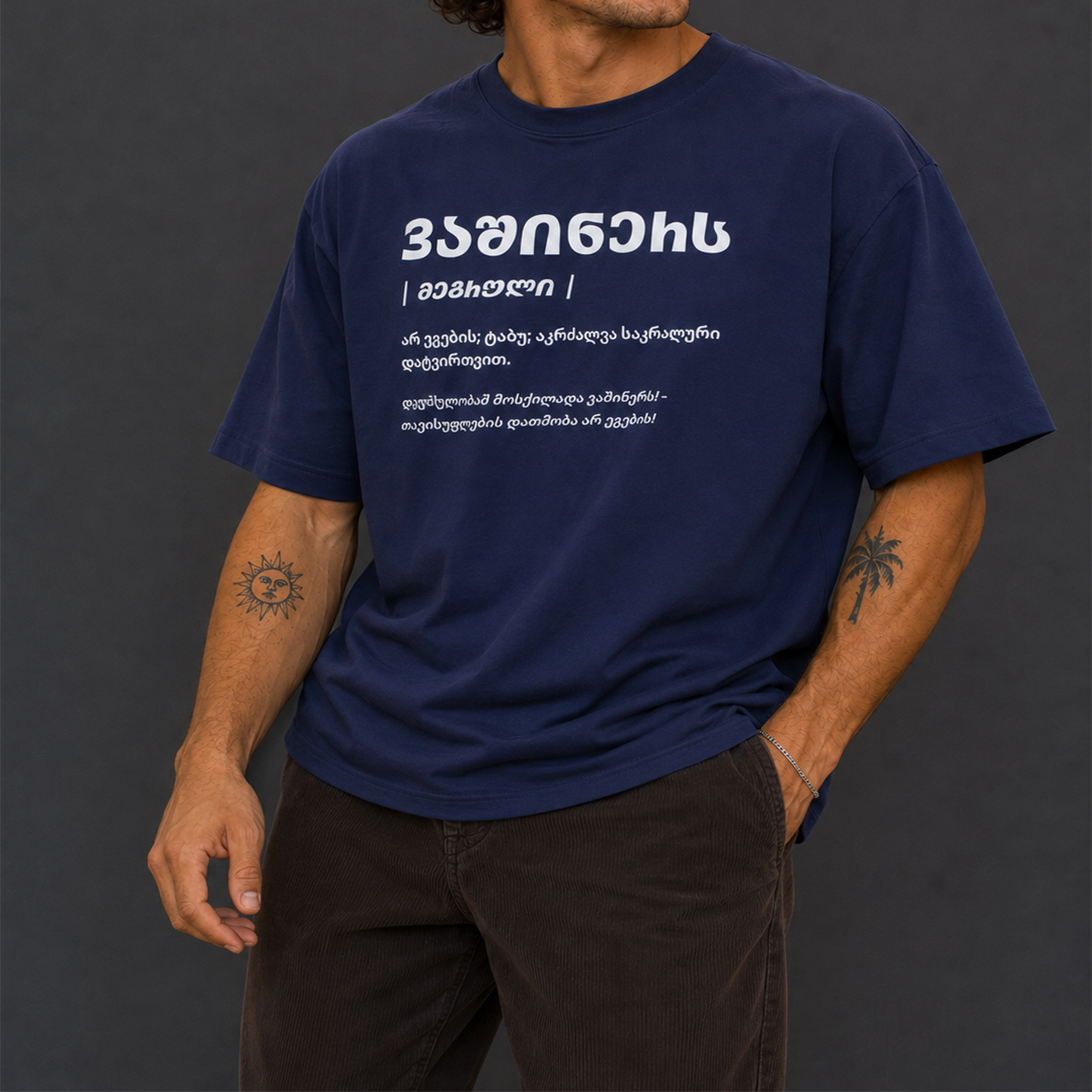 A man wearing a navy blue short-sleeve t-shirt with white Georgian text printed on the chest, paired with dark brown pants, set against a dark gray background.