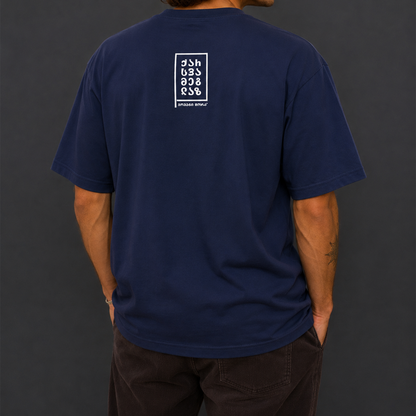 A man viewed from the back wearing a navy blue t-shirt with a white graphic print on the upper back, paired with dark brown pants, set against a dark gray studio background.