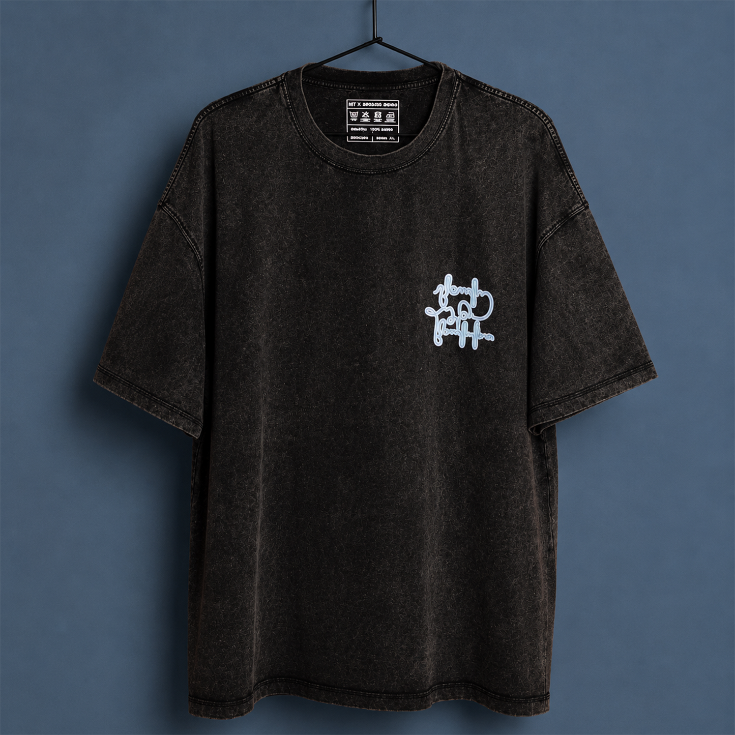 A black acid-wash short-sleeve t-shirt hanging on a black hanger against a slate blue-gray background. The shirt features a small white graffiti-style graphic on the left chest.