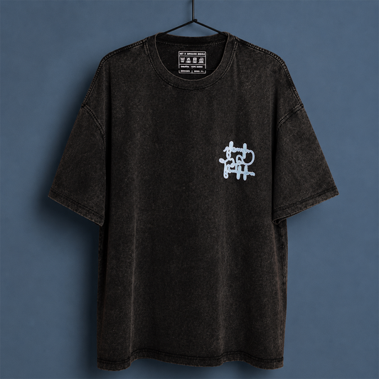 A black acid-wash short-sleeve t-shirt hanging on a black hanger against a slate blue-gray background. The shirt features a small white graffiti-style graphic on the left chest.