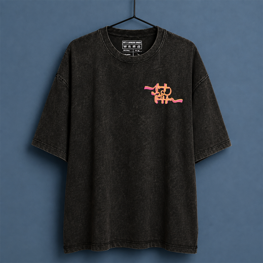 A washed black acid-wash t-shirt hanging on a black hanger against a slate blue background. The shirt features a pink and orange abstract graphic on the left chest resembling a stylized character.