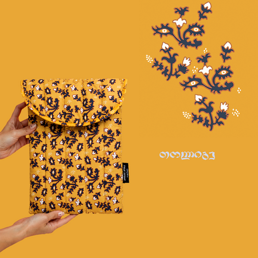 A mustard yellow quilted fabric pouch with a navy and white floral pattern, held by two hands against a solid mustard yellow background. A matching printed card is shown in the upper right corner with stylized text.